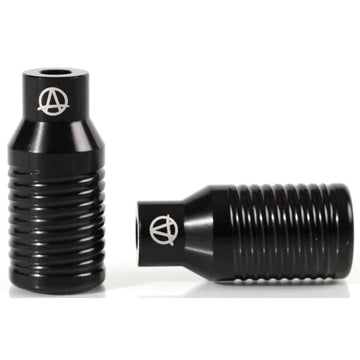 Two Apex Bowie Aluminium Stunt Scooter Pegs in black are displayed, featuring a cylindrical and ribbed design. One peg is placed upright while the other is laid on its side. Both showcase the Apex logo, a stylized "A" within a circle, indicative of their high-quality craftsmanship.